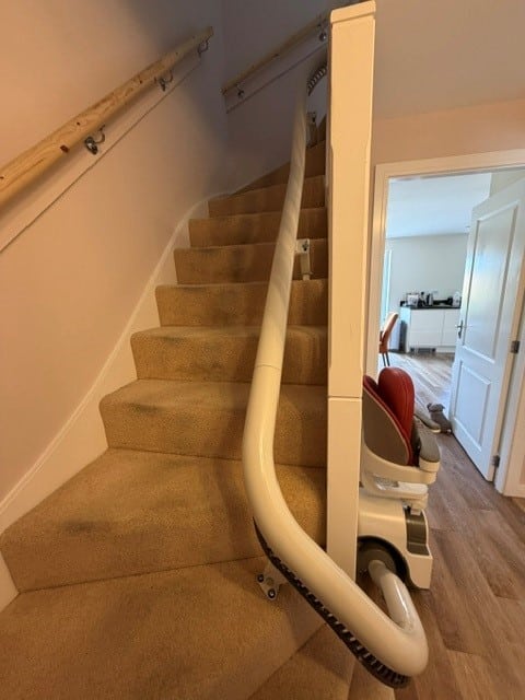 A beige stairlift runs along a carpeted staircase with a white railing and a red-upholstered chair at the bottom, in a home hallway.