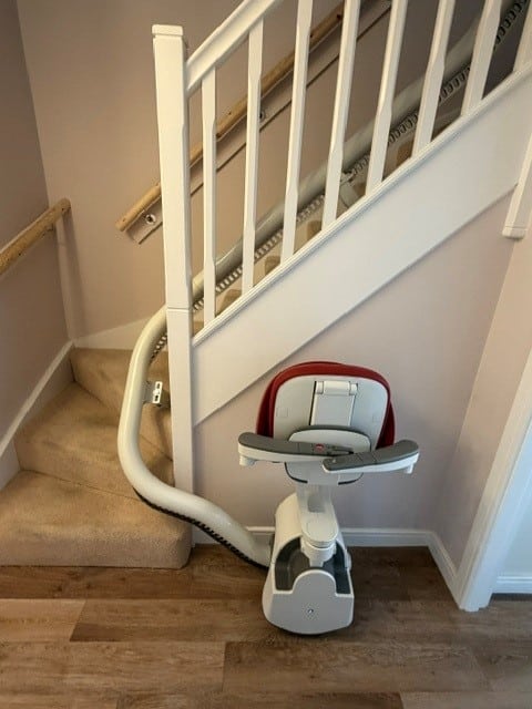 Indoor stairlift chair mounted on a curved staircase, white frame with a red seat cushion, ready to transport a person up or down the stairs.